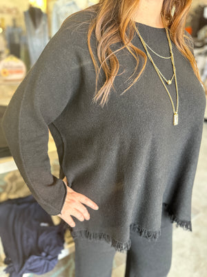 Person wearing a dark gray sweater with fringe details, standing in a blurred indoor setting.