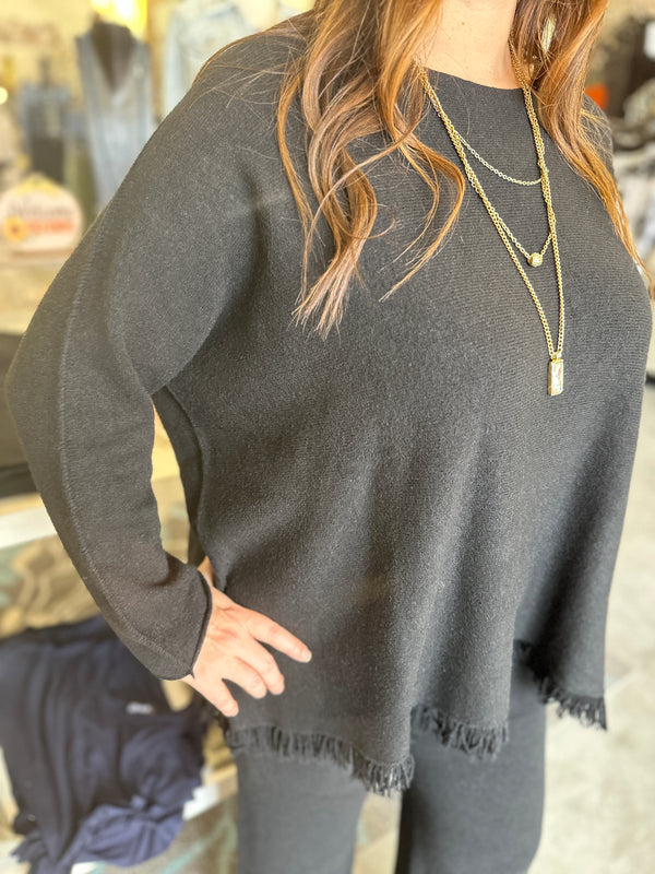 Person wearing a dark gray sweater with fringe details, standing in a blurred indoor setting.